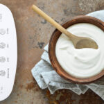 Cremiger, selbst gemachter Joghurt in einer Holzschale mit Holzlöffel, daneben ein KLAMER Brotbackautomat mit aktivem Joghurtprogramm